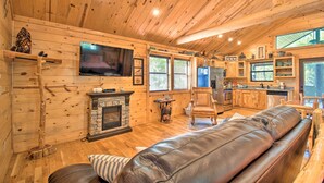 A cozy cabin interior with a stone fireplace, leather sofa, wooden walls, and a kitchen area. - Broken Bow Hideaway w/ Hot Tub & Fire Pit! (Broken Bow)