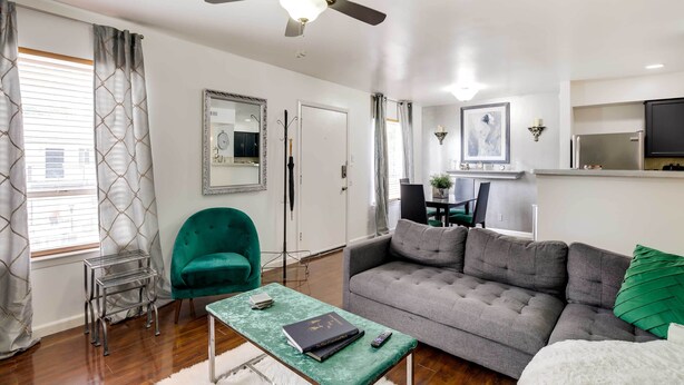 A modern living room with a grey sofa, a green armchair, and a small coffee table. There is a ceiling fan, a dining area in the background, and a kitchen with a TV. - Chic Dallas Condo w/ Pool, Walk to Knox Street! (Dallas)
