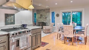 Een moderne keuken met een stenen open haard, een eetgedeelte met gestreept beklede stoelen en een groot raam met uitzicht op groen.