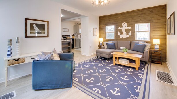 A living room with a blue sofa, a wooden coffee table, and a patterned rug. There is a wooden wall with an anchor decoration and a window with blinds. - Walk to Lake: Nautical Akron Cottage w/ Deck! (Akron)