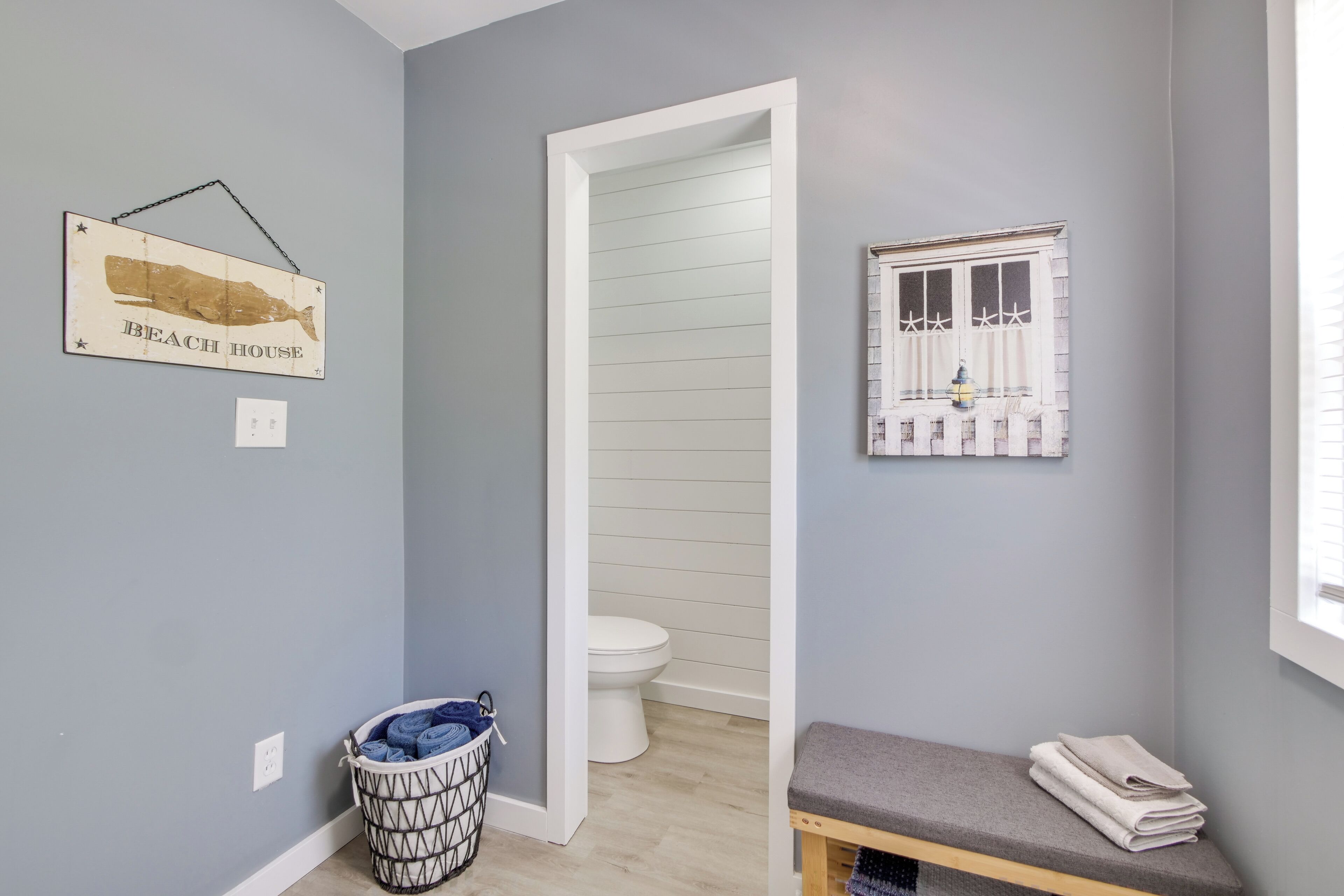 Une salle de bain comprenant des toilettes, un banc, un tableau accroché au mur et un panier à linge.