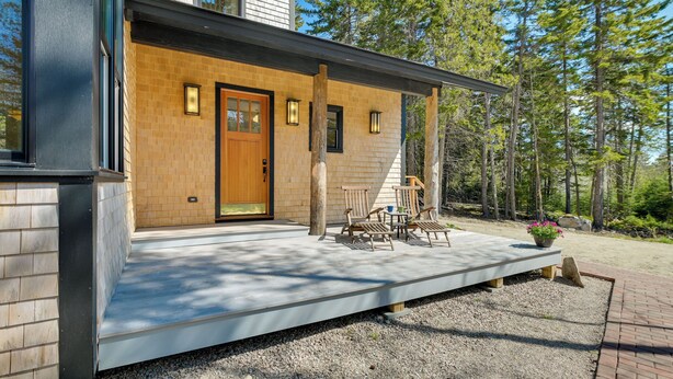Une maison moderne avec une porte d’entrée en bois, un porche couvert doté de meubles d’extérieur et une allée en gravier.