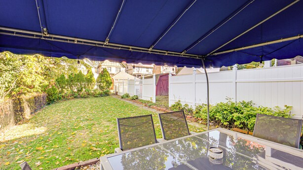 Eine überdachte Terrasse im Freien mit einem Glastisch und Stühlen, umgeben von einem weißen Zaun und Pflanzen.