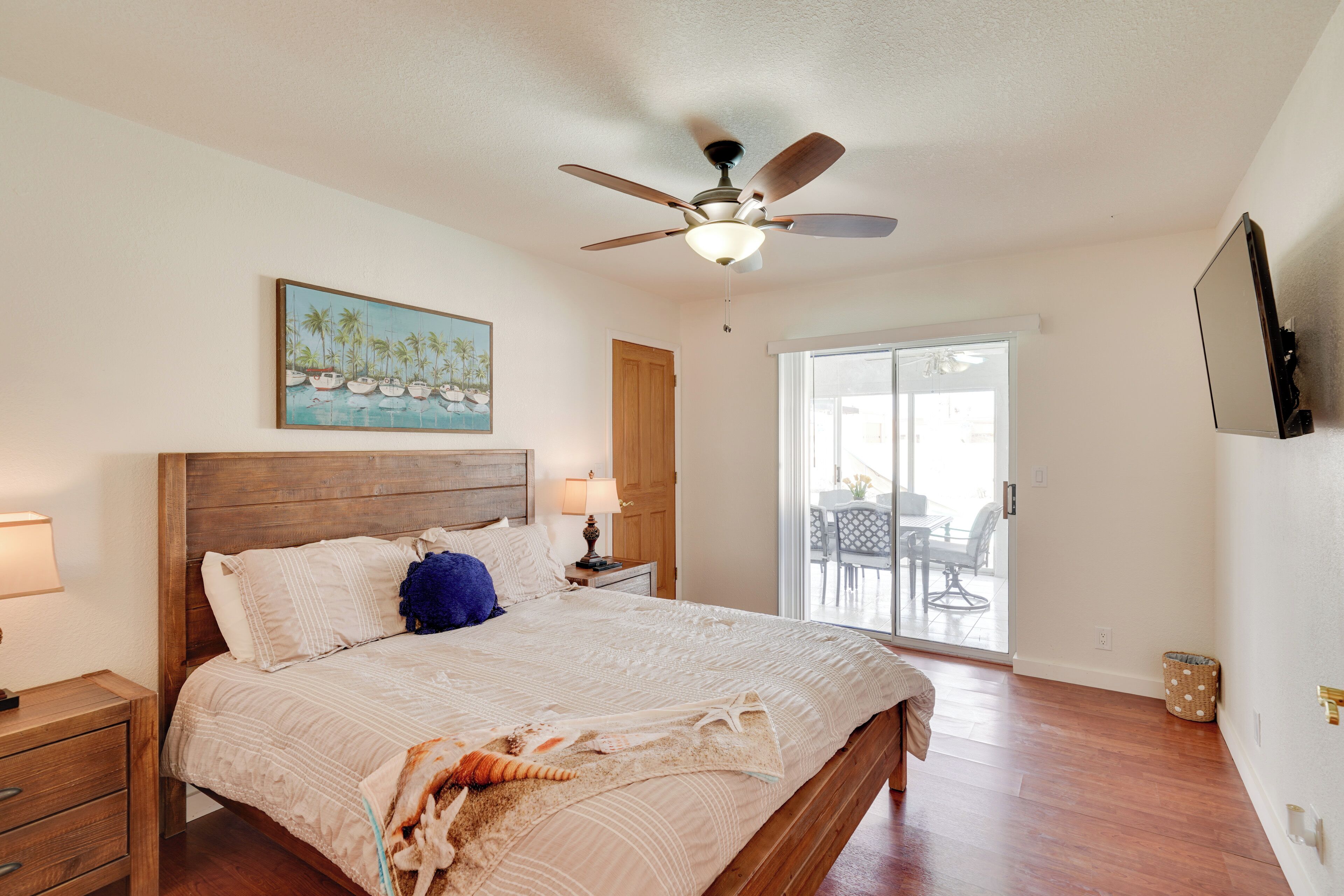 A bedroom with a wooden bed, a ceiling fan, a painting of boats, and a sliding glass door to a balcony.
