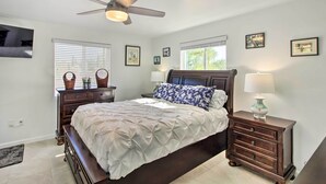 Quarto com cama, mesa de cabeceira, cômoda, ventilador de teto e janela com persianas.