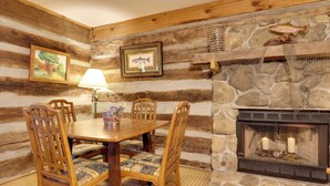 A rustic wooden dining area with a stone fireplace, wooden chairs, and a table with a lamp. - Fire Pit & Deck! Cozy Smoky Mtn Retreat on River (Townsend)
