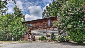 A rustic log cabin with a wooden porch, surrounded by lush greenery and flowering bushes. - Fire Pit & Deck! Cozy Smoky Mtn Retreat on River (Townsend)
