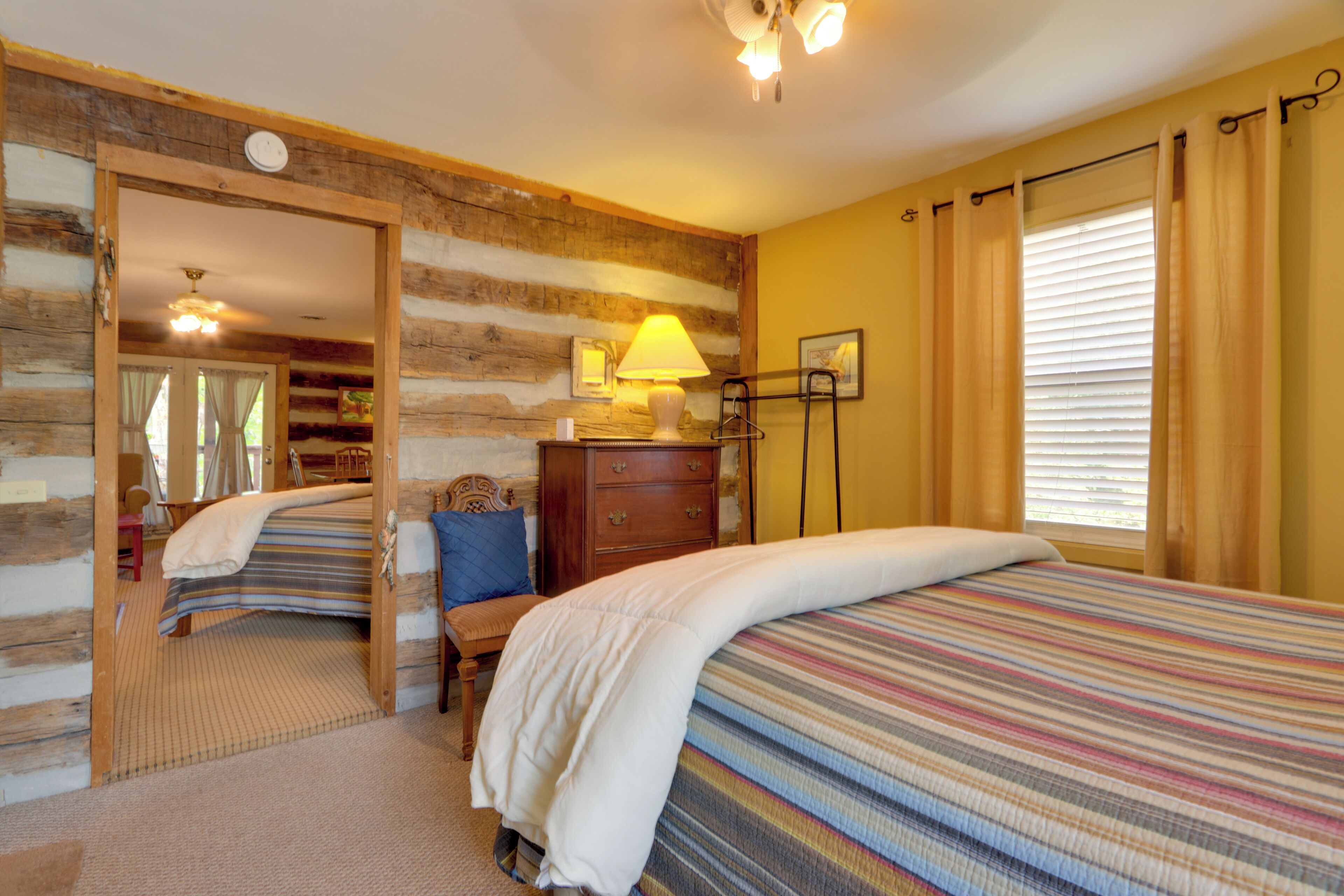 A cozy bedroom with a wooden log wall, a bed with a striped bedspread, a bedside lamp, a dresser, and a window with curtains.
