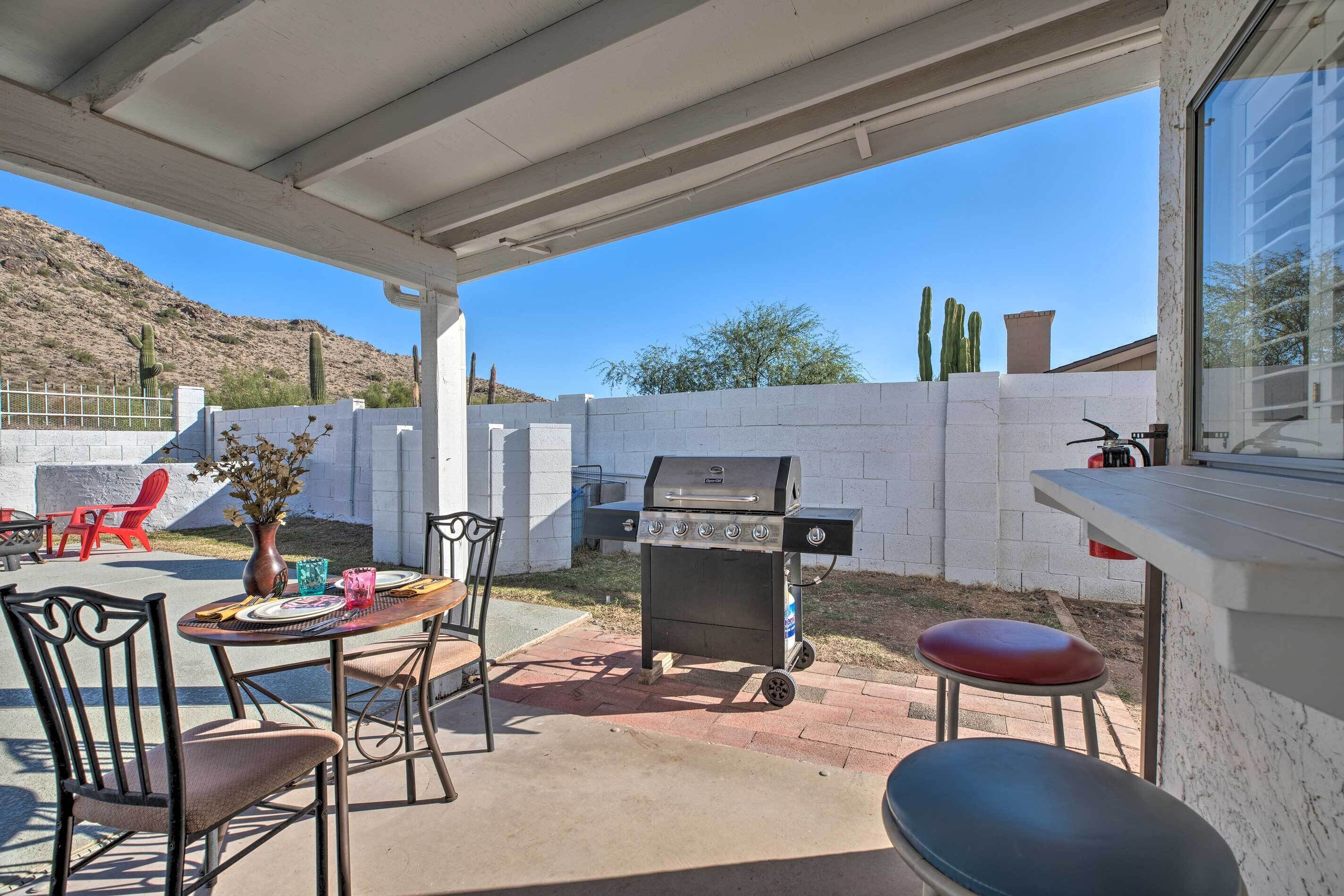 An outdoor patio with a barbecue grill, a table with chairs, and a window overlooking a desert landscape.