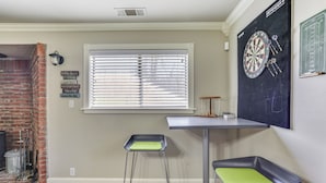 Een kamer met een metselwerk openhaard, een raam met jaloezieën, een dartbord en twee stoelen.