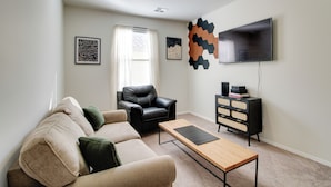 Ein Wohnzimmer mit einem beigefarbenen Sofa, einem schwarzen Ledersessel, einem Holztisch, einer schwarzen TV-Konsole und einem Fenster mit weißen Vorhängen.