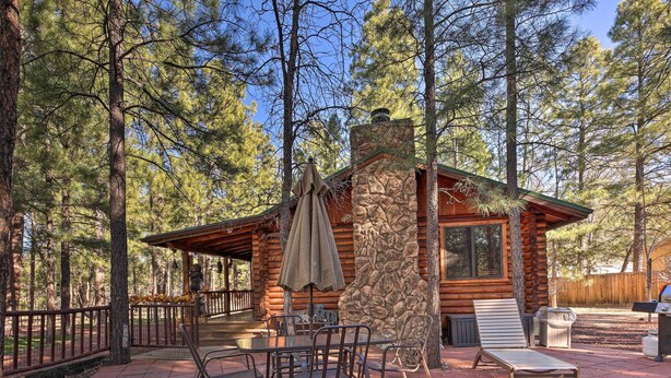 A log cabin with a stone chimney, outdoor seating area with metal chairs and a table, and a covered patio with an umbrella. - Family & Dog Friendly: Rustic Lakeside Cabin (Lakeside)
