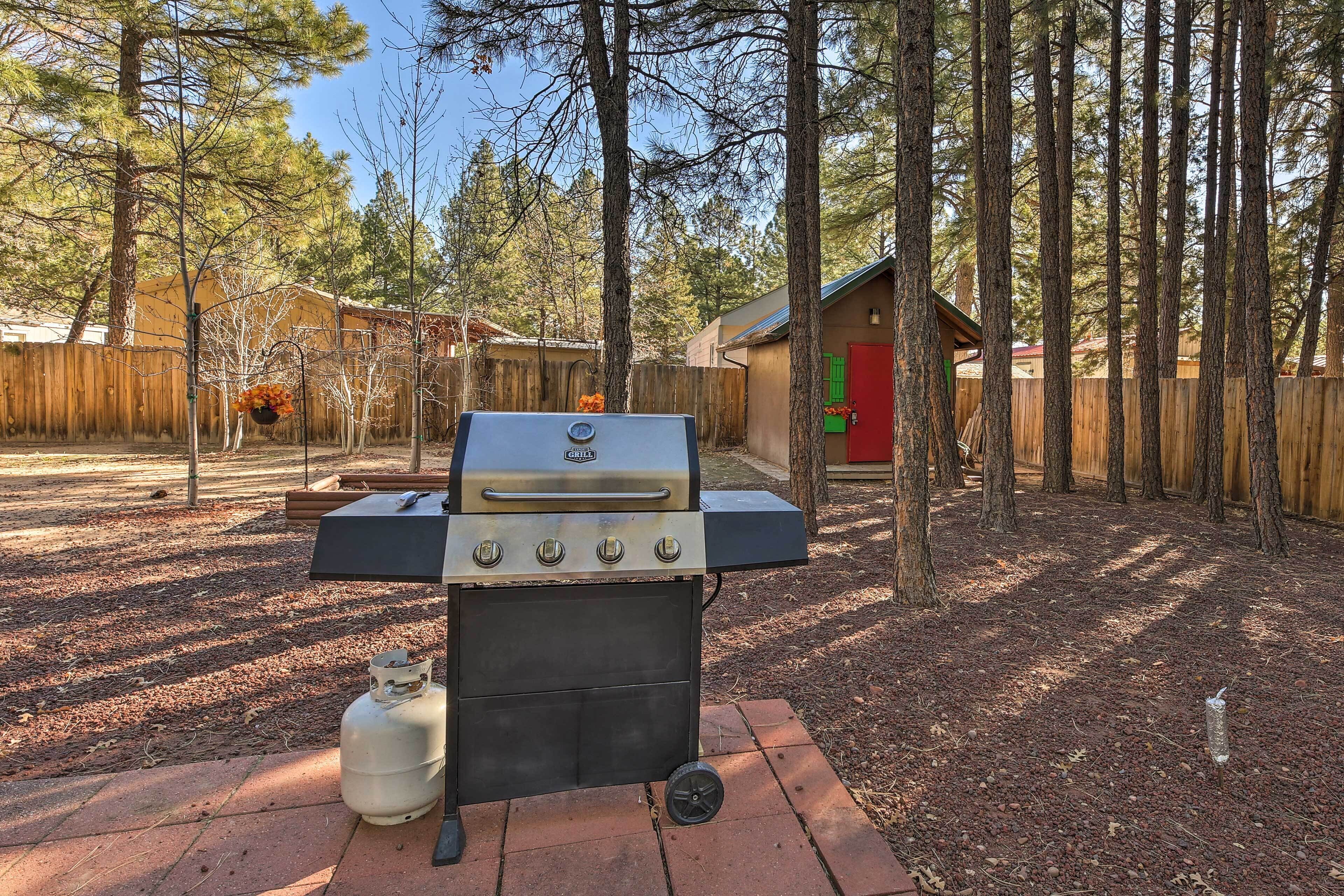 A barbecue grill with a propane tank, set on a brick patio surrounded by trees.