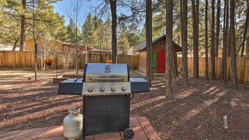 A barbecue grill with a propane tank, set on a brick patio surrounded by trees.