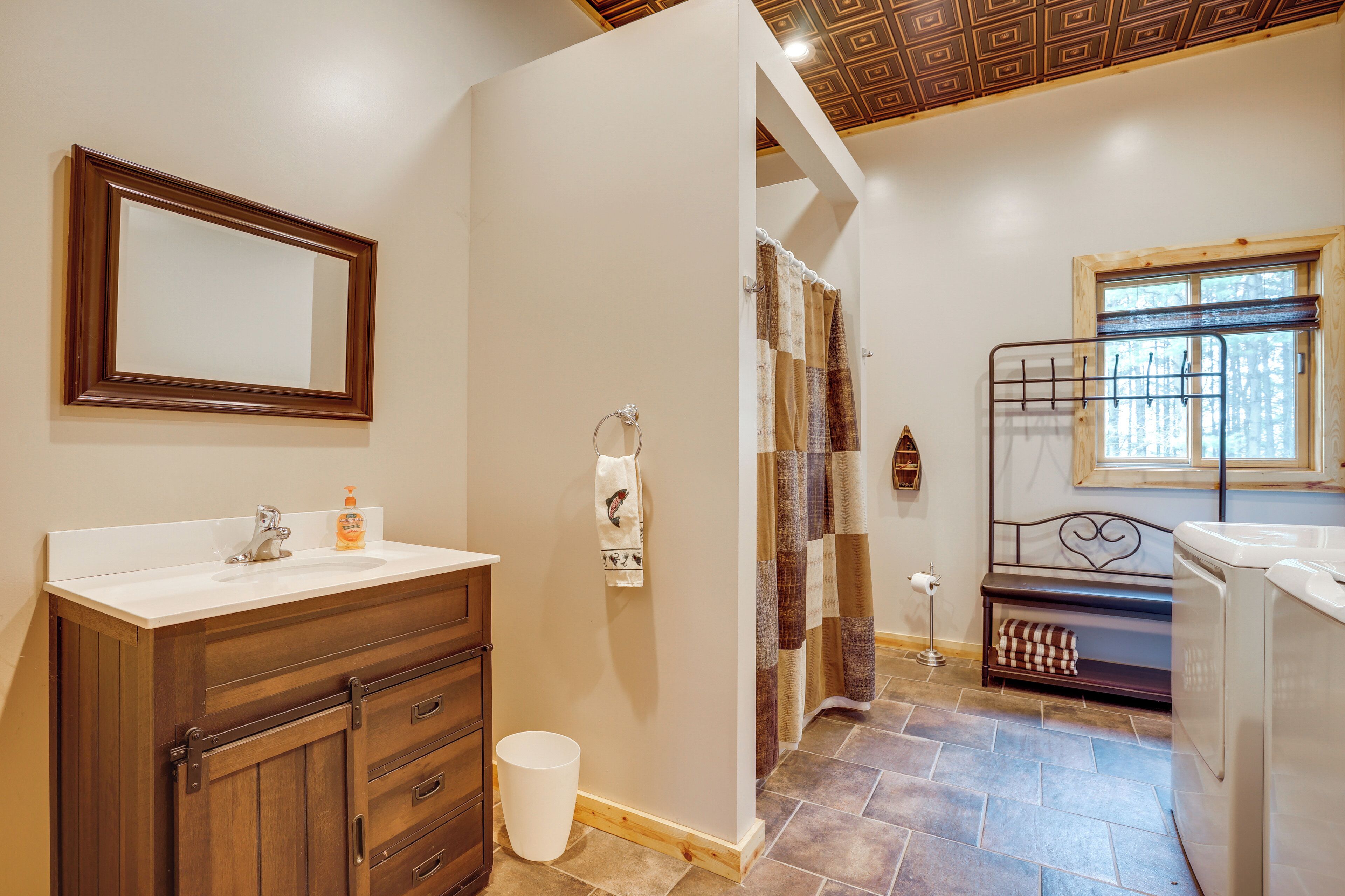 A bathroom with a wooden vanity, a mirror, a sink, a shower with a quilted curtain, a bench with a heart-shaped metal frame, and a window with a view of trees.
