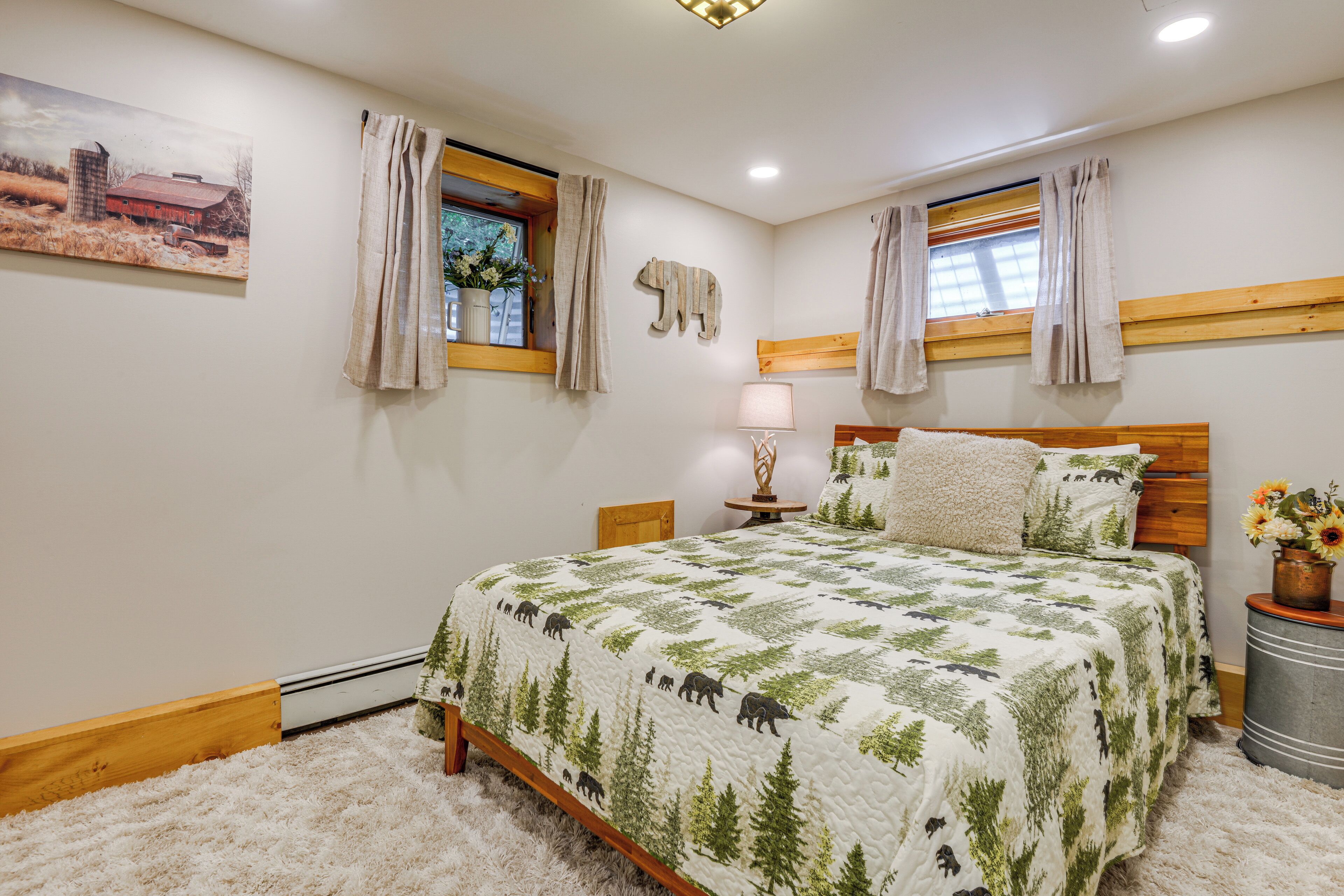 A bedroom with a bed, a nightstand, a lamp, a window with curtains, a wall-mounted bear decoration, and a painting of a barn.