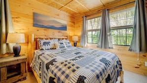 Ein Schlafzimmer einer Holzhütte mit einem großen Bett, zwei Nachttischlampen, einem Gemälde an der Wand und einem Fenster mit Vorhängen.