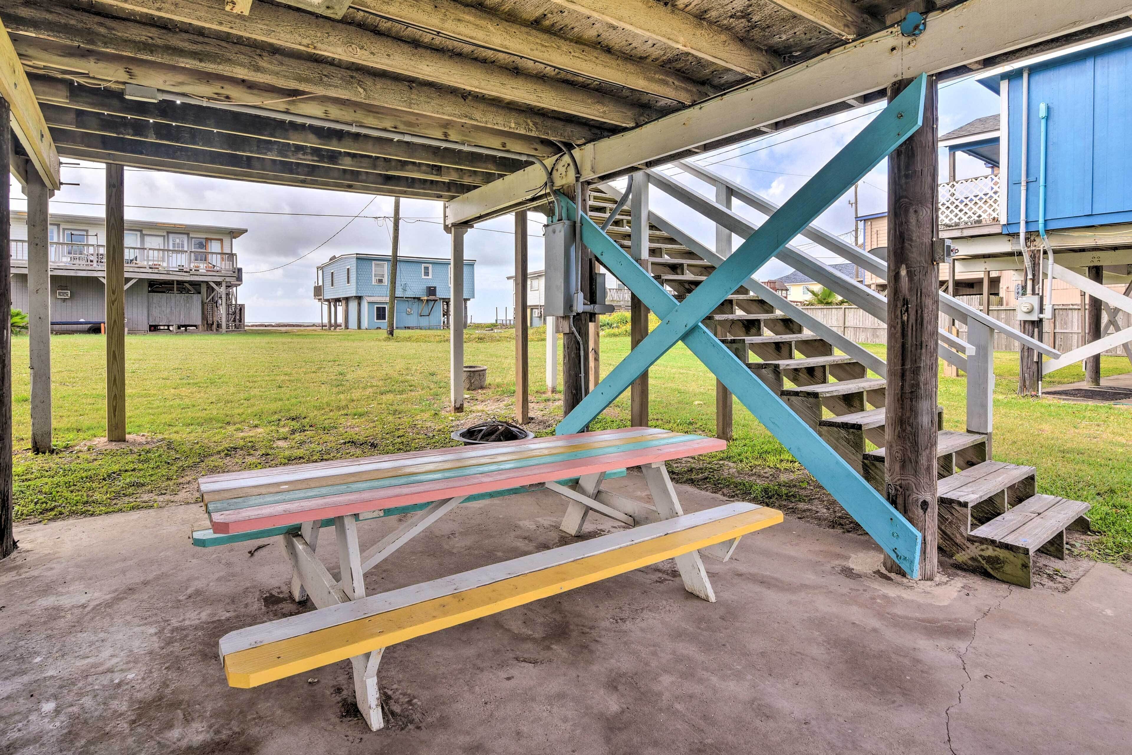 Una mesa de picnic bajo una zona cubierta, con una escalera que lleva a un edificio azul.