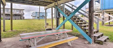 Una mesa de picnic bajo una zona cubierta, con una escalera que lleva a un edificio azul.