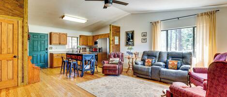 A spacious living area with a sofa, a coffee table, and a dining table with chairs. There is a ceiling fan, wooden flooring, and a view of the outdoors through a window.