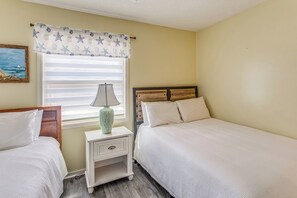 A bedroom with two beds, a nightstand with a lamp, a window with blinds, and a painting on the wall. - Myrtle Beach Cottage, Walk to Pools + Beach! (Myrtle Beach)