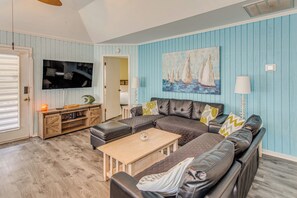 A living room with a sectional sofa, a coffee table, a flat-screen TV, and a painting of sailboats.