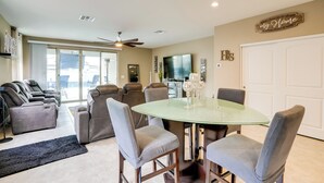 A modern living room with a glass-top dining table, gray upholstered chairs, a ceiling fan, and a large TV. - Private Pool: Relaxing Phoenix Area Vacation Home! (Buckeye)