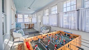 A spacious room with a foosball table, wicker furniture, and large windows with sheer curtains.