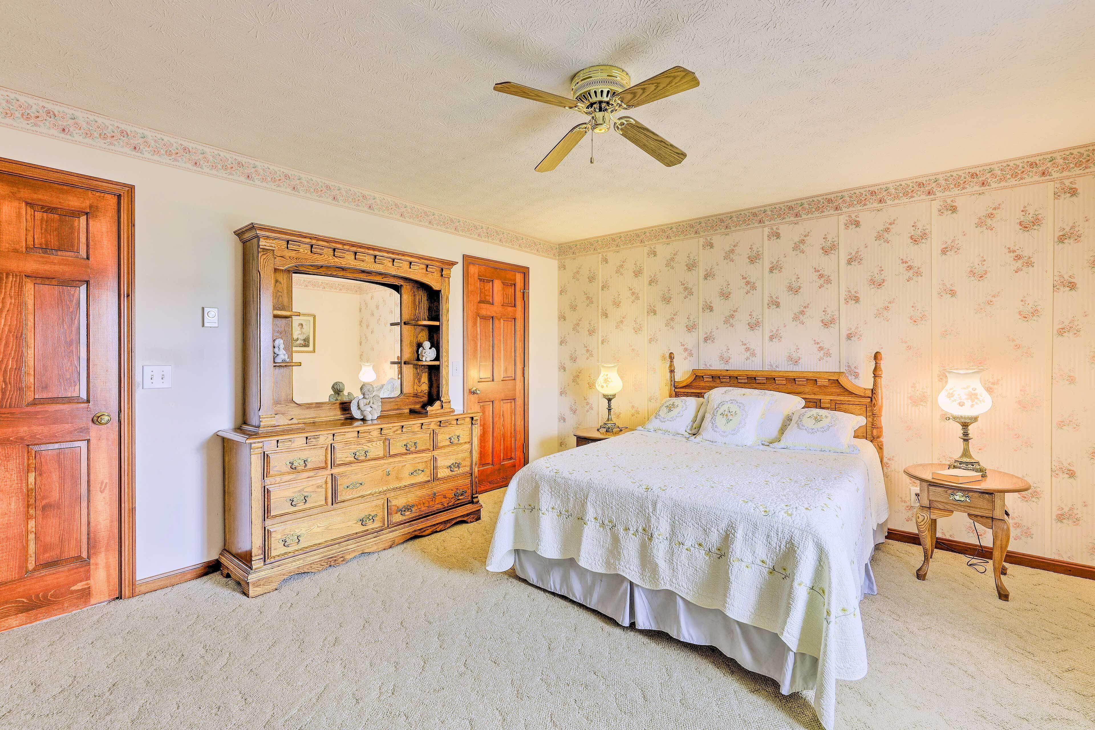 A bedroom with a wooden dresser, a large mirror, a bed with white bedding, a ceiling fan, and floral wallpaper.