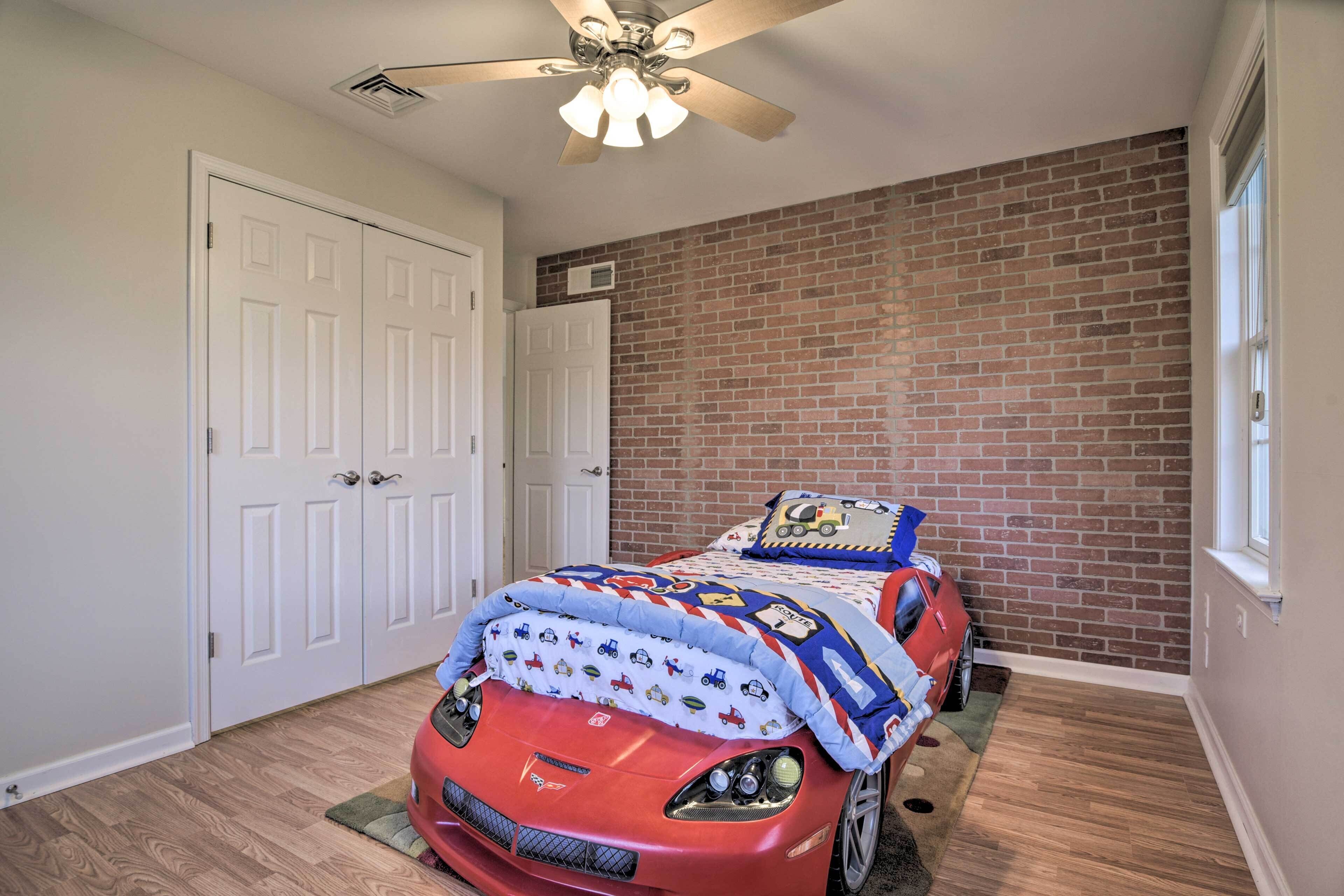 Un dormitorio con pared de ladrillo, ventilador de techo, cama con cubrecama con temática de autos y un auto de juguete rojo.