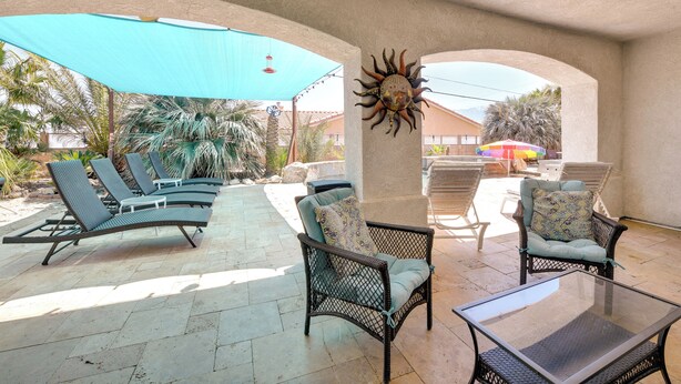 Een overdekt terras met lounge fauteuils en een glazen tafel, met een decoratieve zon aan de muur en een blauwe parasol.