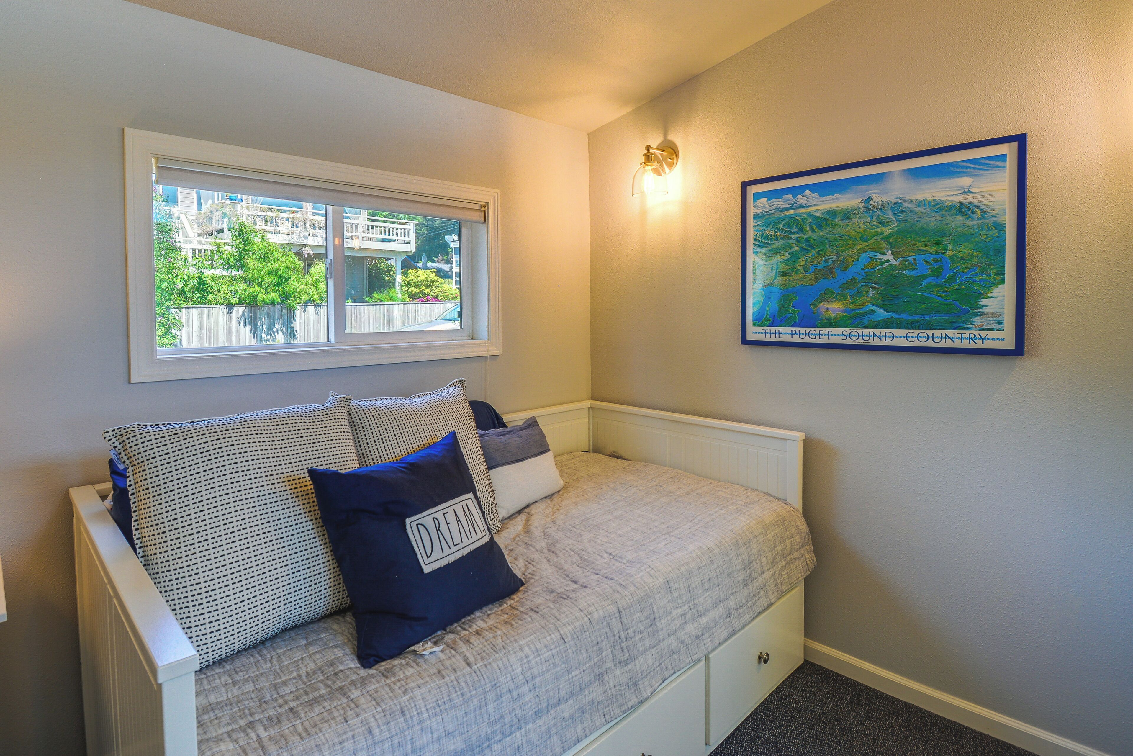 Une chambre à coucher avec un lit, une fenêtre, une lampe fixée au mur et un tableau encadré.
