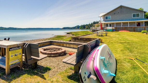 A lakeside property with a wooden deck, a covered seating area, and a large house with a porch. - Waterfront Hood Canal Cottage: Steps to the Beach! (Belfair)
