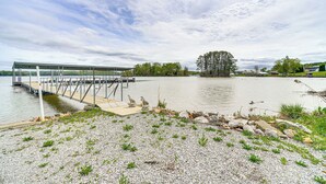 Een uitzicht op het meer, met een steiger die het water in reikt, omgeven door kiezelstenen en schaarse begroeiing.