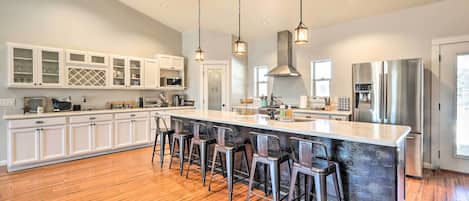 A modern kitchen with a large island, bar stools, white cabinetry, stainless steel appliances, and pendant lighting.