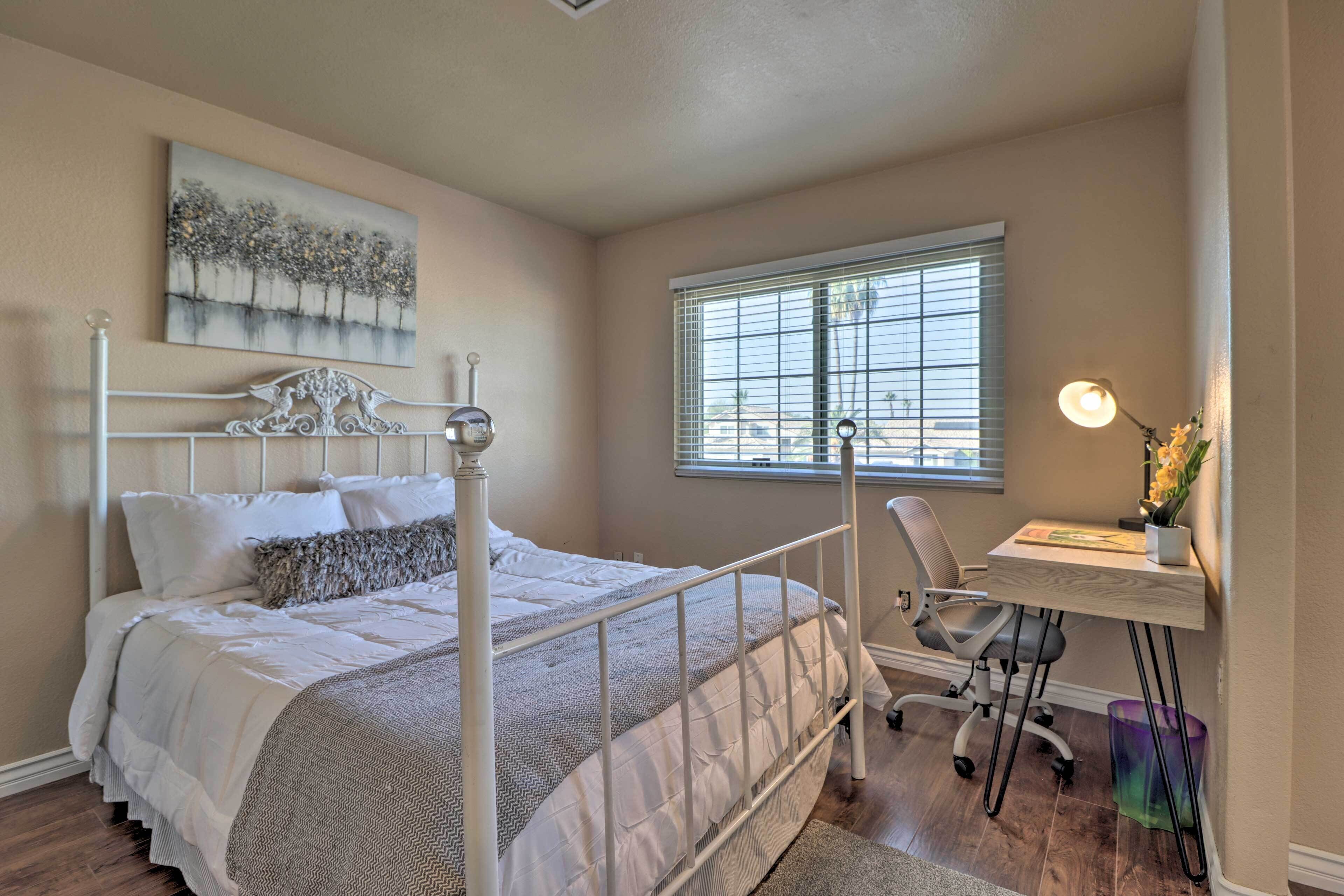 A bedroom with a four-poster bed, a desk with a chair, a window with blinds, and a painting on the wall.