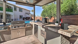 Une cuisine extérieure équipée d’un barbecue, d’un évier et de placards de rangement, donnant sur un jardin avec une piscine et des meubles de patio.