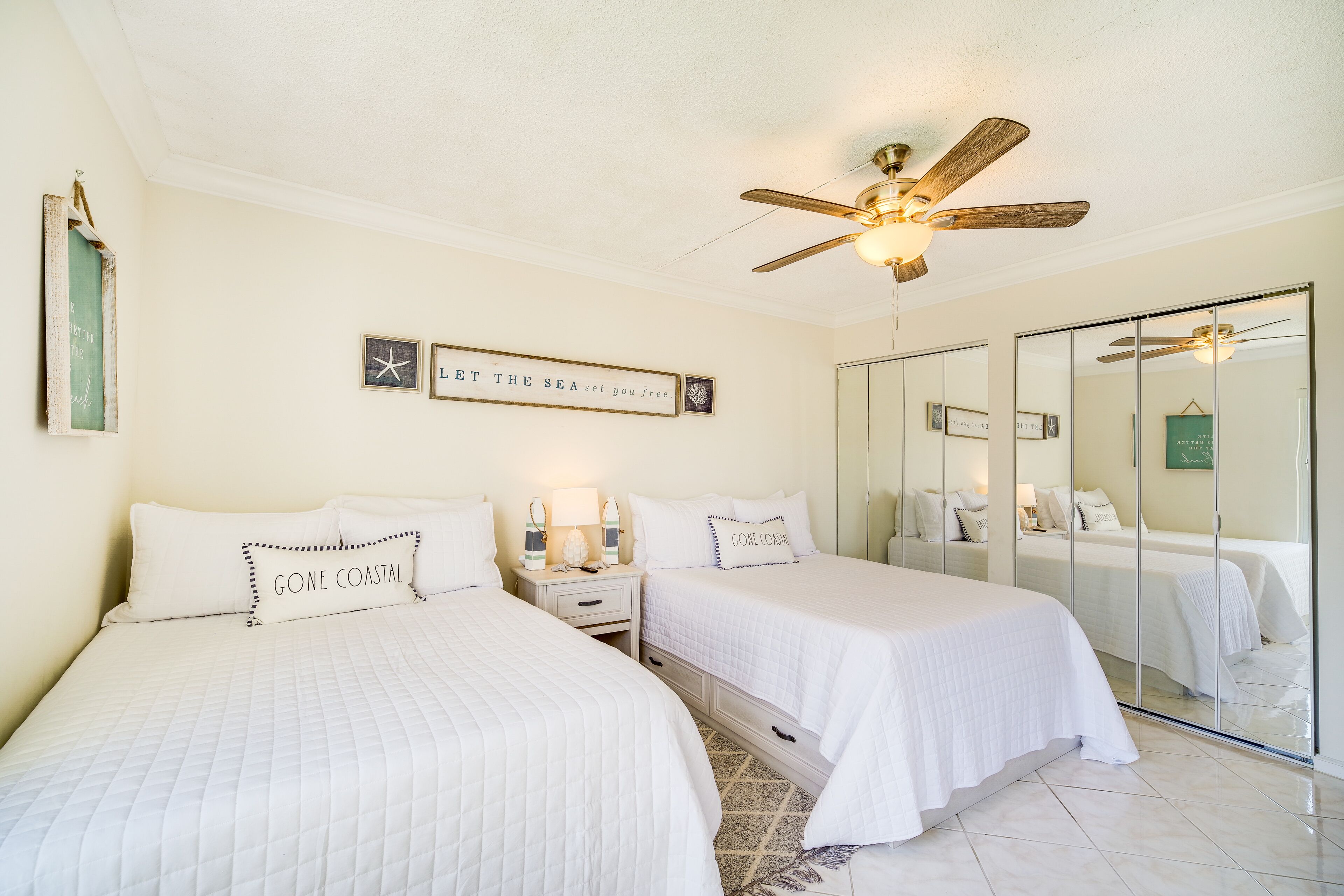 A hotel room with two beds, a ceiling fan, a mirror, and a wall with framed pictures and a sign.