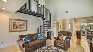 A spacious living room with a brown leather sofa, a brown leather armchair, and a patterned rug. A black spiral staircase is visible in the background.