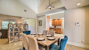 A dining area with a wooden table set for four, surrounded by chairs. There is a bookshelf with decorative items and a ceiling fan above.