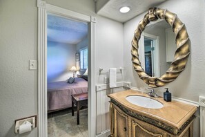 A bathroom with a beige vanity, a large mirror with a decorative frame, and a view into a bedroom with a bed and a nightstand.