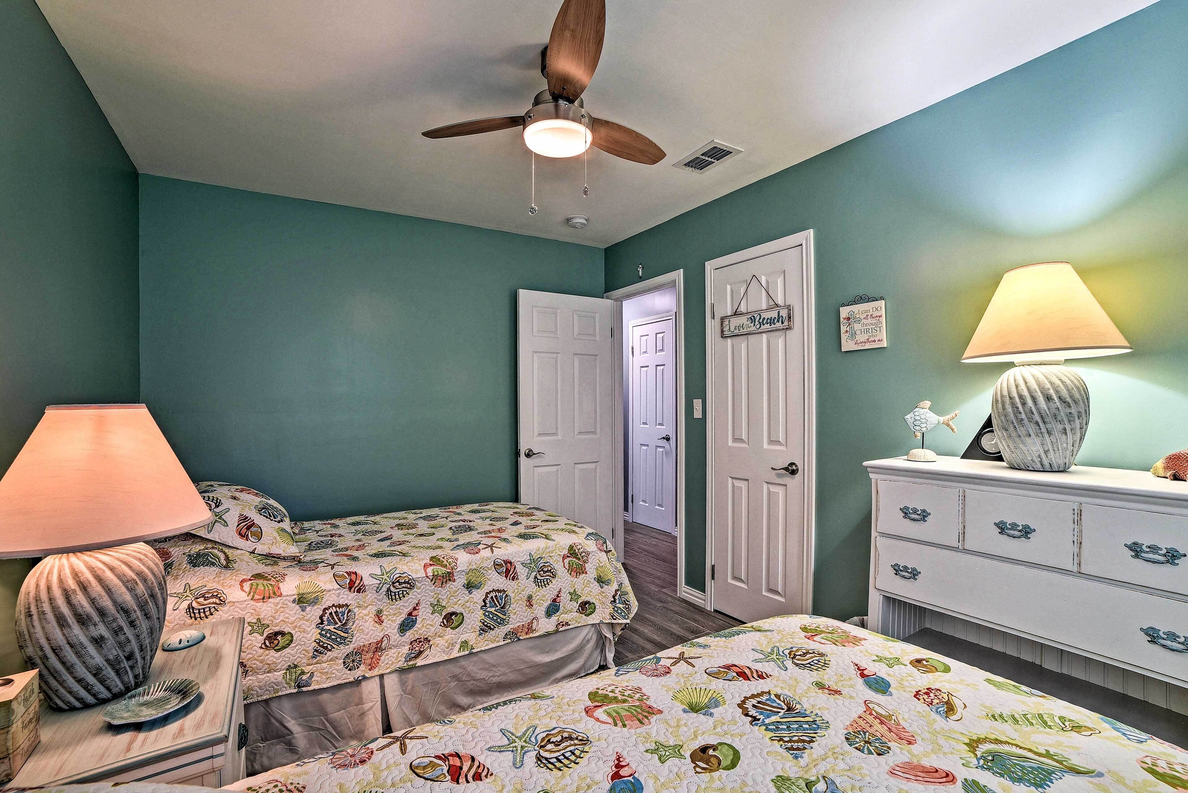 A bedroom with two beds, a ceiling fan, a lamp, a dresser, and a door leading to another room.