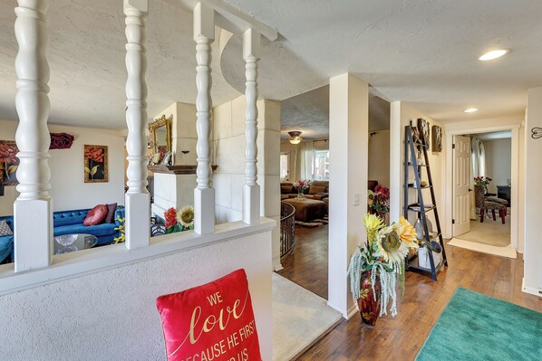 Amplia sala de estar con sofá azul, almohadones decorativos y un cojín rojo con un mensaje romántico, además de una escalera con barandilla blanca que conduce a otra habitación.