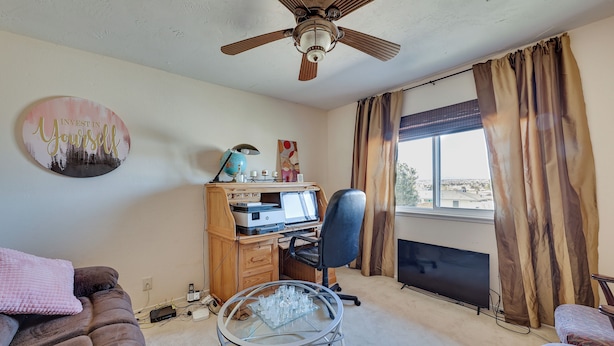 Une pièce avec un ventilateur de plafond, un bureau avec un ordinateur, une chaise, une table en verre avec des tasses, un canapé et une fenêtre avec des rideaux.