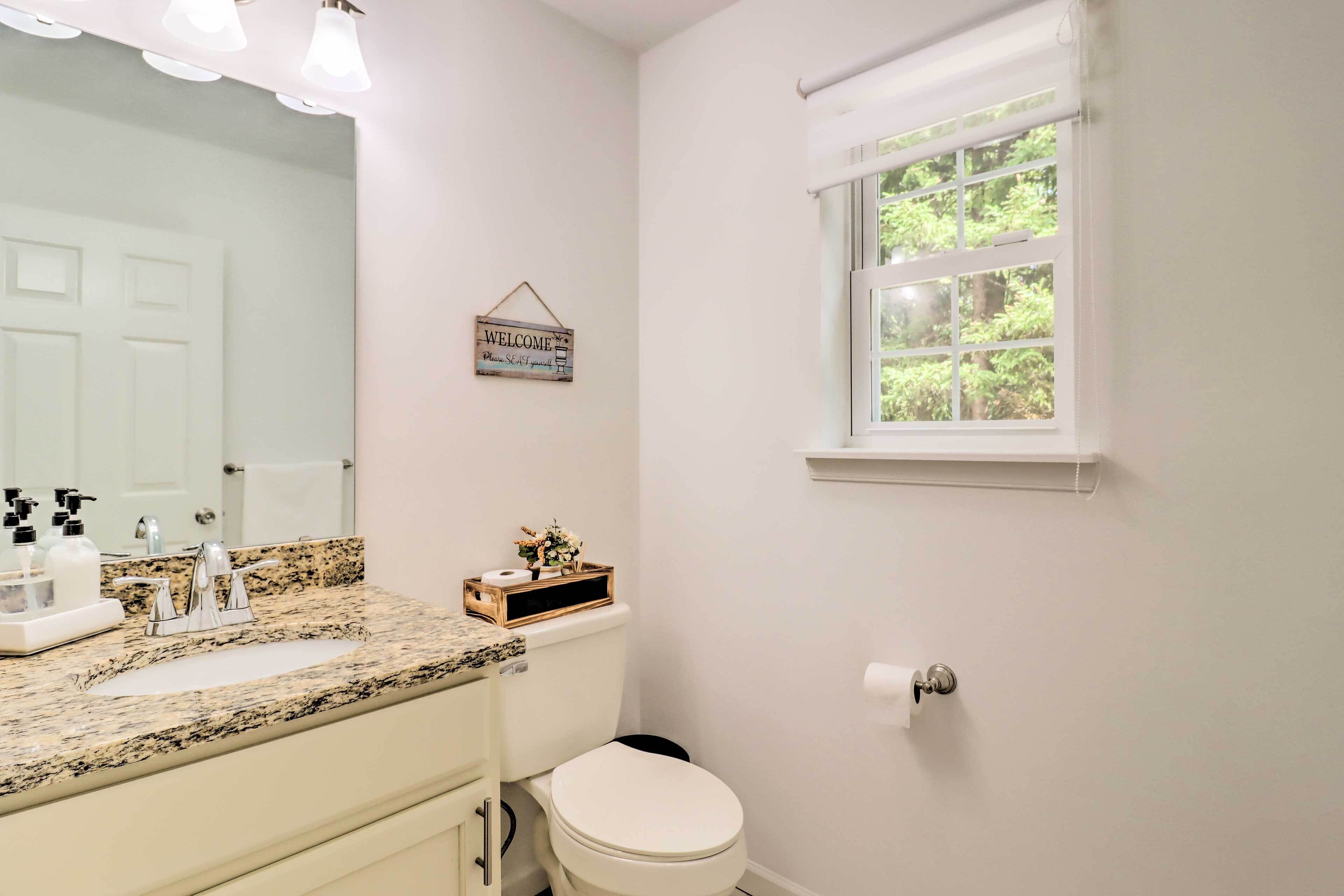 A bathroom with a marble countertop, a toilet, a sink with soap dispensers, a mirror, and a window with a view of greenery.