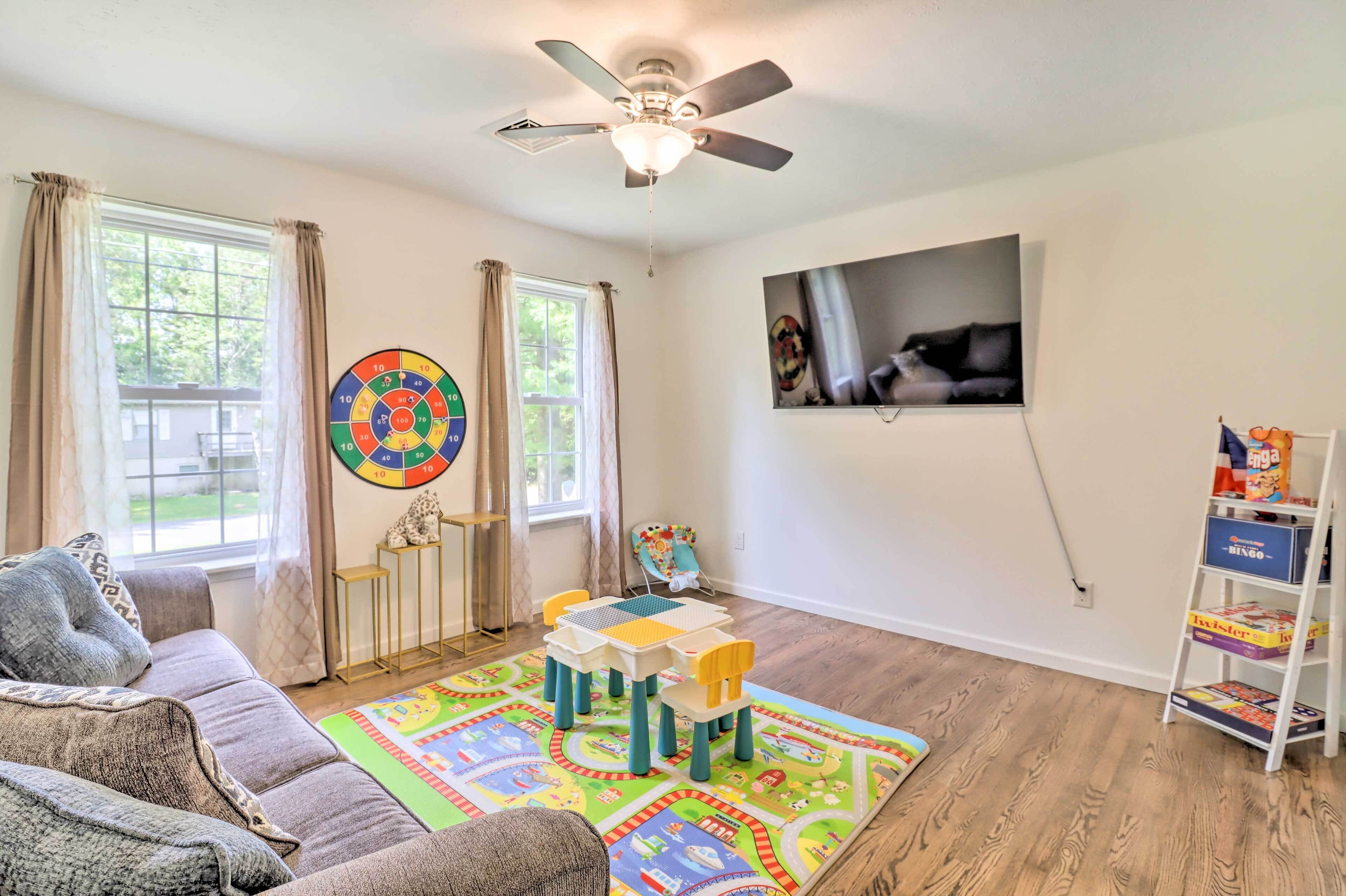 A living room with a ceiling fan, a colorful play mat, a sofa, a TV mounted on the wall, and a bookshelf with children's books.
