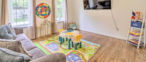 A living room with a ceiling fan, a colorful play mat, a sofa, a TV mounted on the wall, and a bookshelf with children's books.