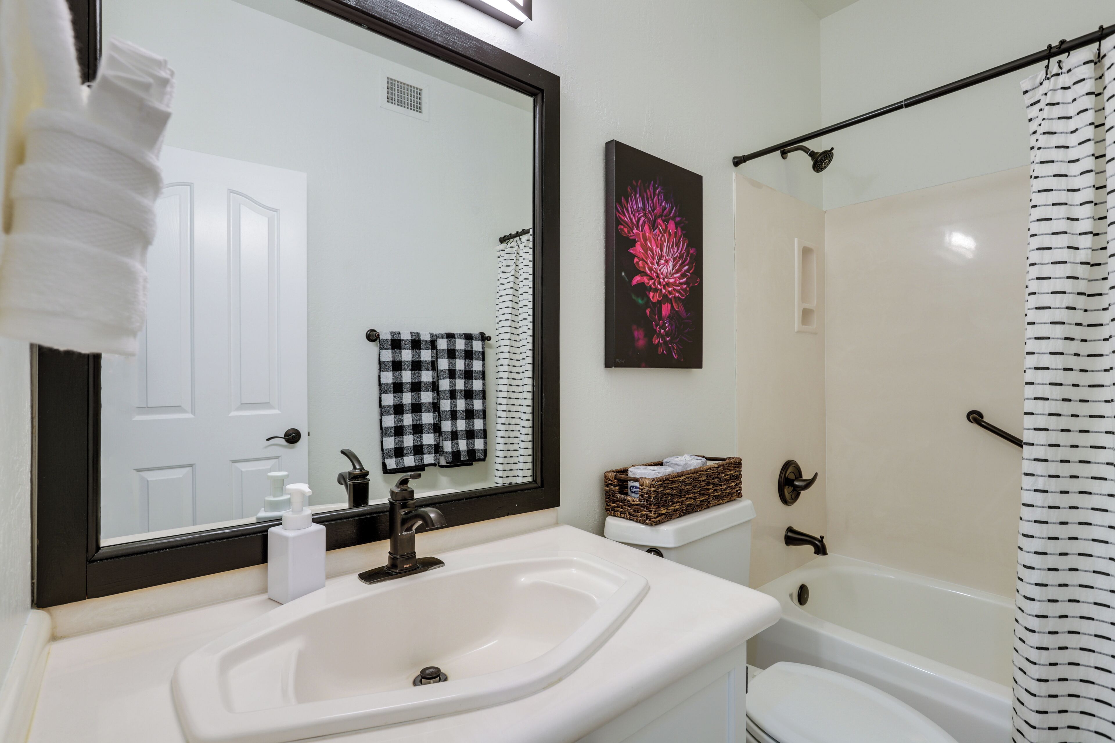Une salle de bain avec un lavabo blanc, un miroir, une serviette à carreaux noirs et blancs, un panier et un rideau de douche.