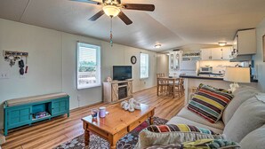A living room with a wooden coffee table, a teal bench, a sofa with colorful pillows, a ceiling fan, and a kitchen in the background. - Peaceful Supply House ~ 2 Mi to Holden Beach! (Supply)