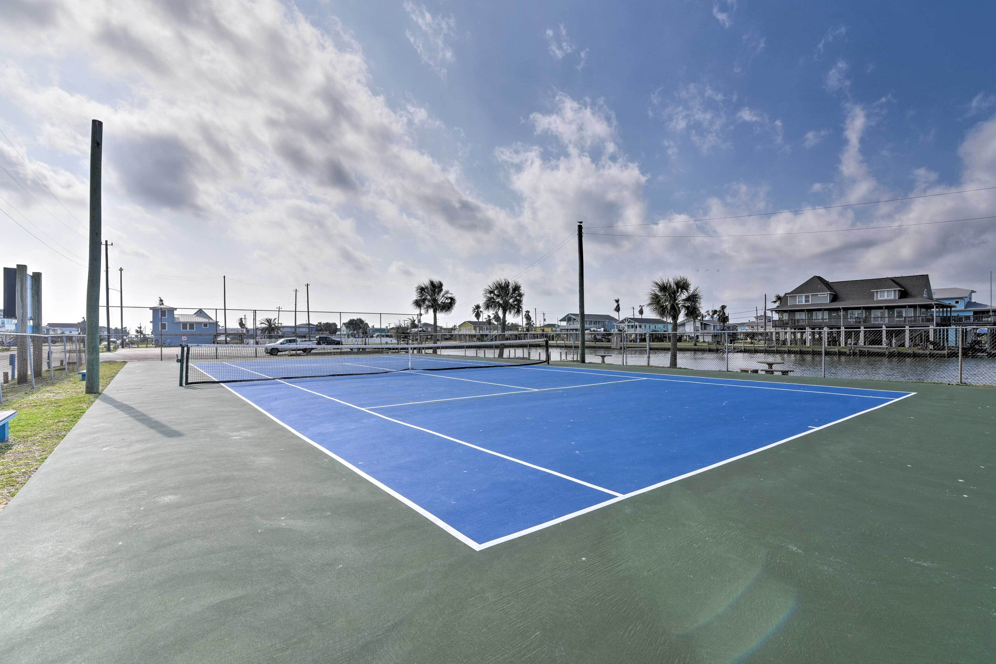 Een tennisbaan met een net, omgeven door een hek, en een waterpartij op de achtergrond.